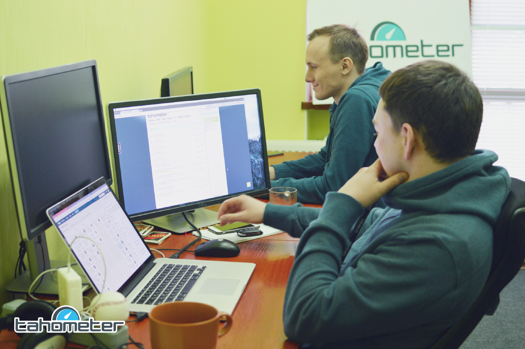 Two young men in teal hoodies working at computers in a bright office with Tahometer branding.