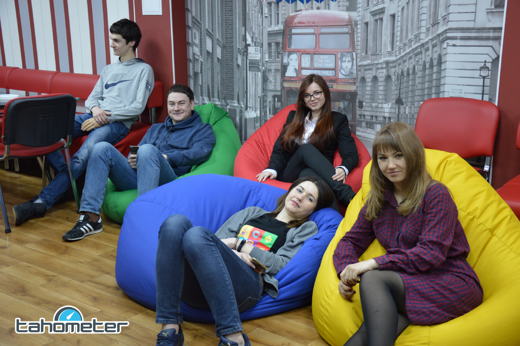Six young people sitting on colorful bean bags in a modern office lounge with London street scene backdrop.